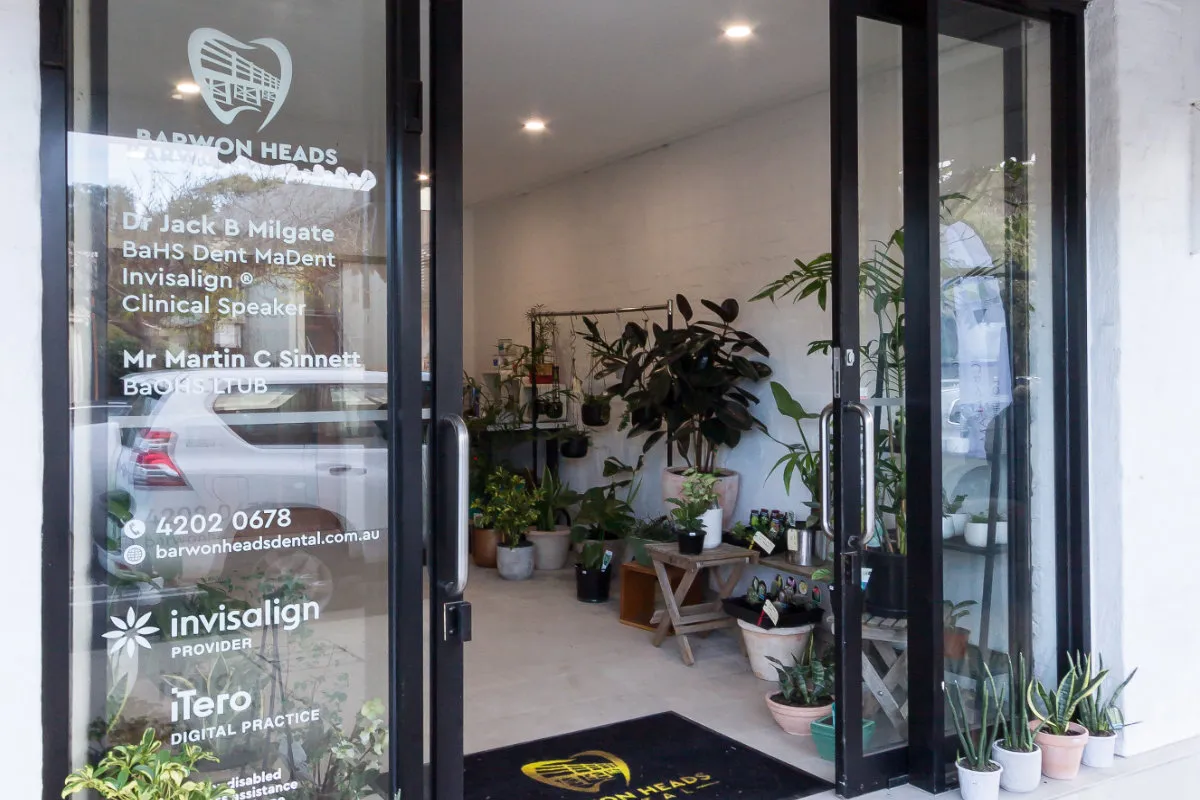 Open glass front doors of Barwon Heads Dental with signage and an indoor plant display on a timber bench.
