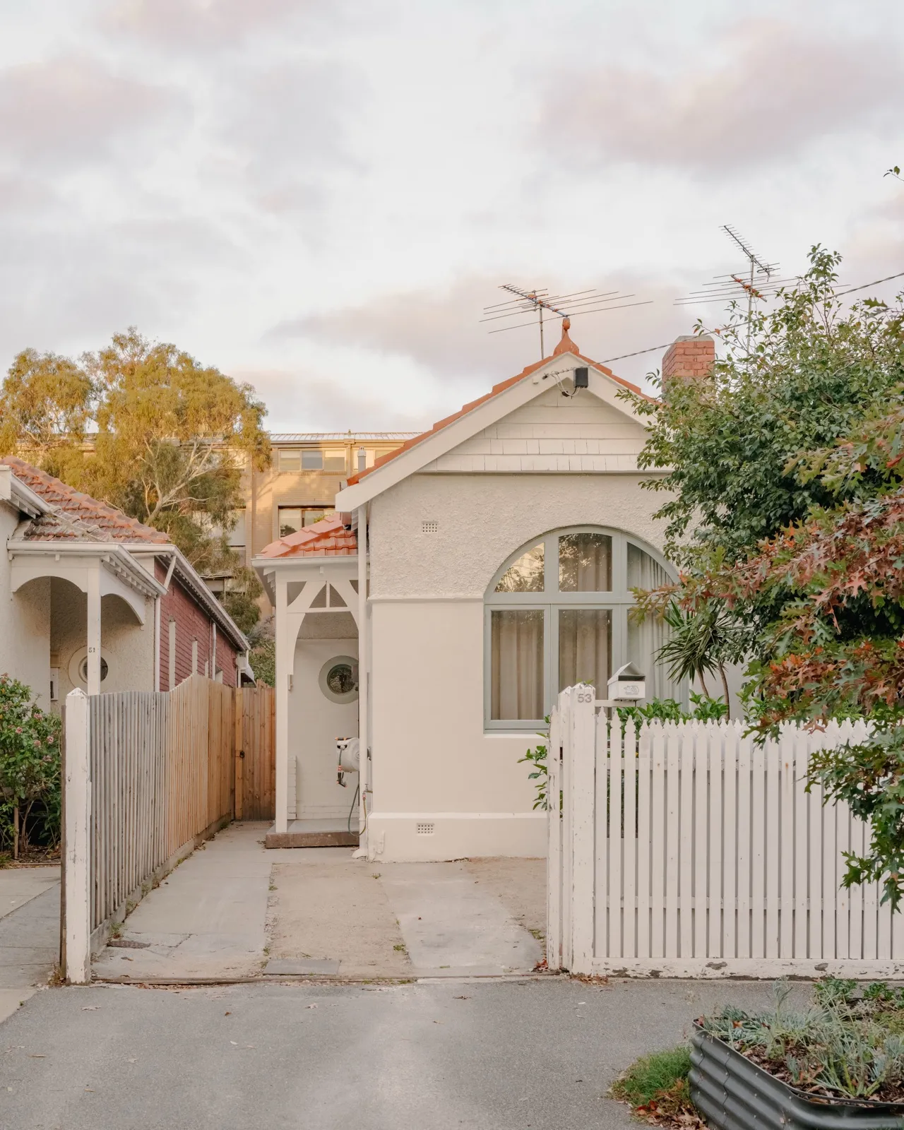 Street view of the white-rendered Edwardian cottage with arched window, terracotta tiled roof, and white picket gate at dusk.