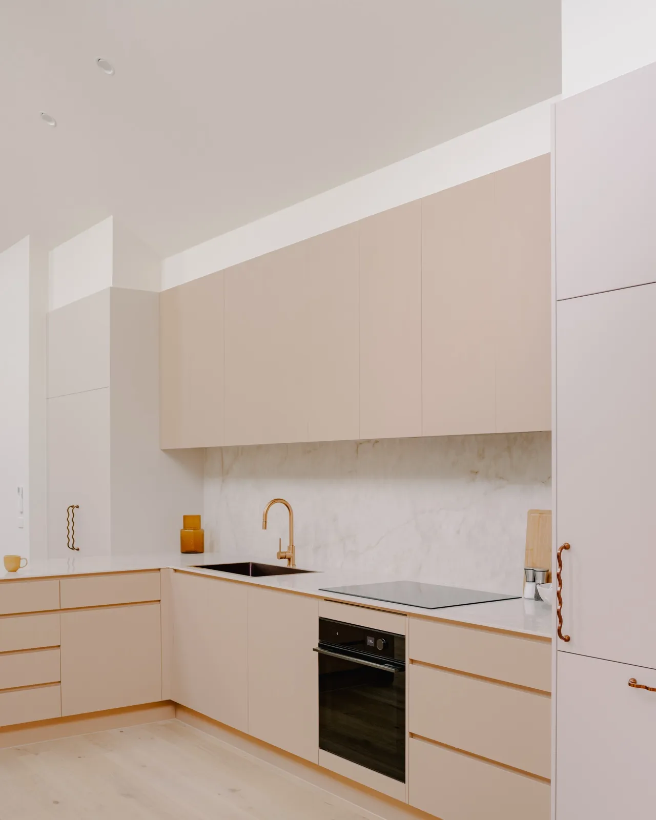 Blush kitchen with handle-less cabinets, marble splashback, brushed copper gooseneck mixer, induction cooktop, and integrated oven.