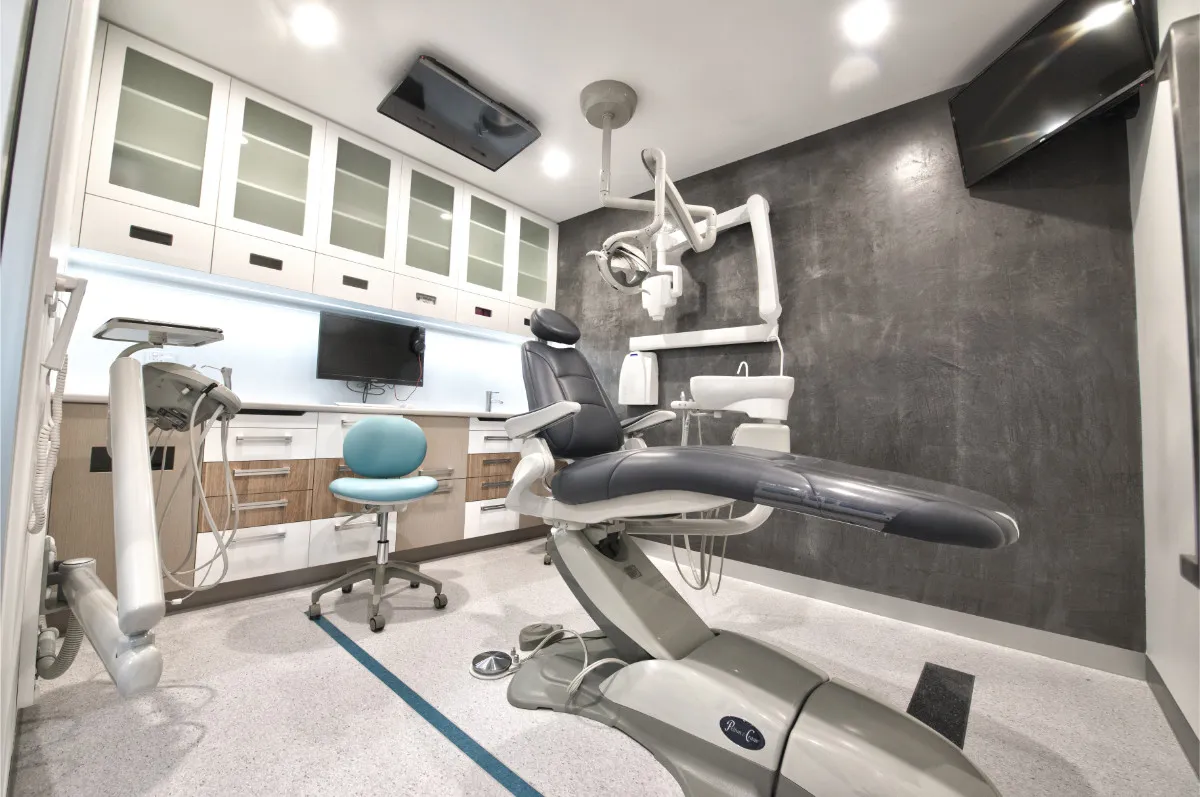 Dental surgery with treatment chair, overhead light, timber cabinetry, and grey concrete-finish feature wall.
