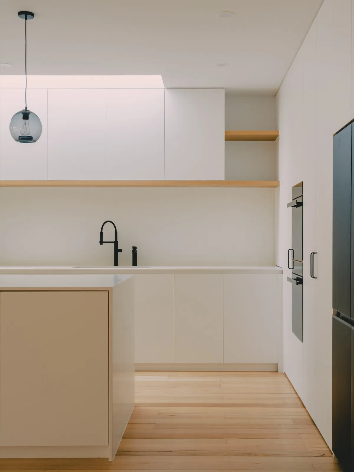 Galley kitchen view with a single smoke-glass pendant, black mixer tap, white cabinets, and integrated wall ovens.