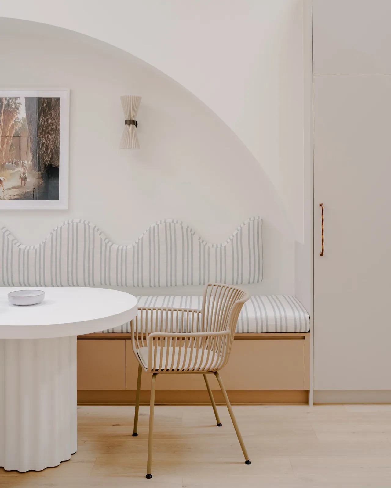 Dining nook with white fluted pedestal table, rattan armchair, and a striped scalloped banquette under a pleated wall sconce.