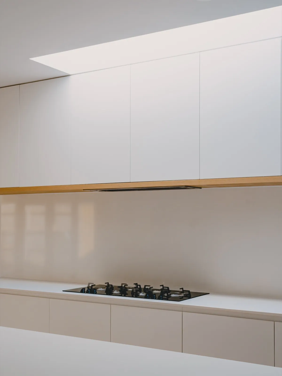 Kitchen detail with a skylight above white overhead cabinets, timber underside trim, and a gas cooktop on the white stone bench.