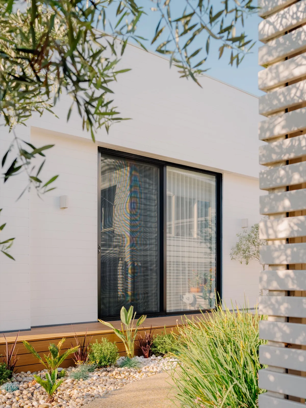 Entry to the home with black-framed sliding doors, white cladding, timber slat screen, and pebble garden bed.