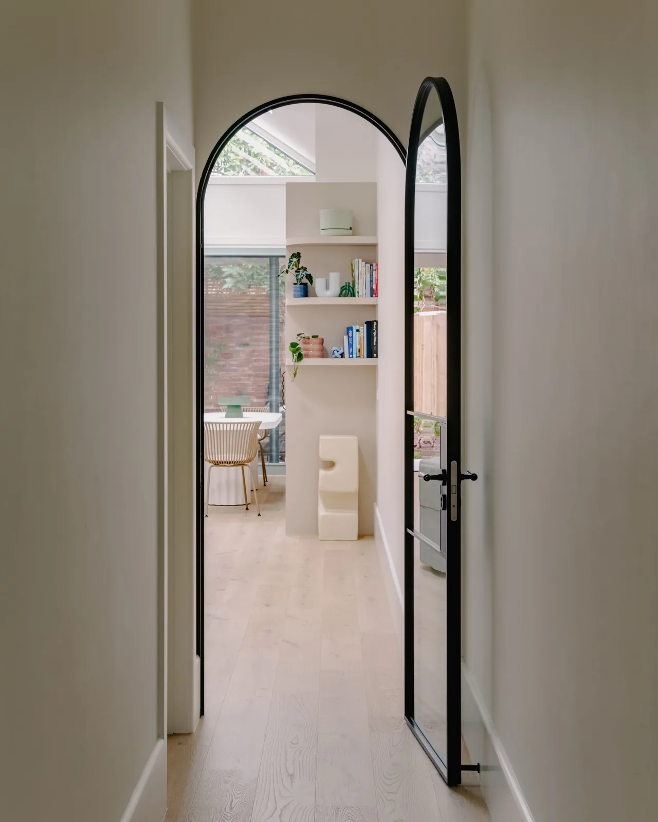 Hallway view through an open black-framed arched door to the rear living space with built-in shelving and a courtyard beyond.