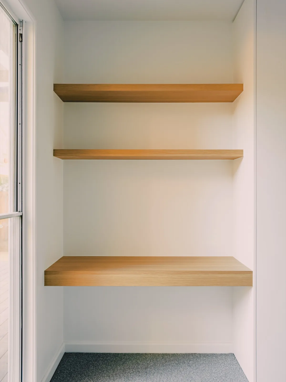 Linen alcove with three floating timber shelves, white walls, and grey carpet flooring beside a window.
