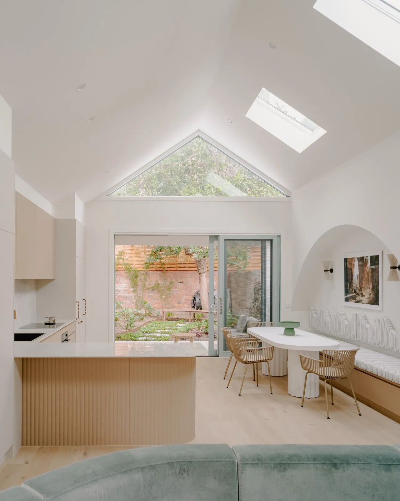 Open kitchen, dining, and living with vaulted ceiling, triangular gable window, striped banquette, and sliding doors to a brick-walled courtyard.