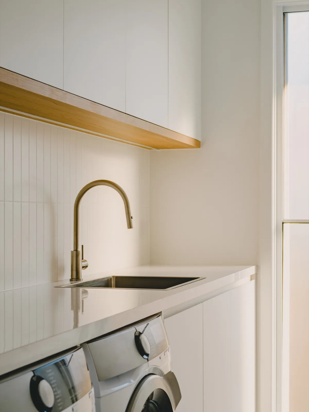 Laundry with vertical-panelled splashback, white cabinets, brass gooseneck mixer, timber underside shelf, and a front-loader washer.