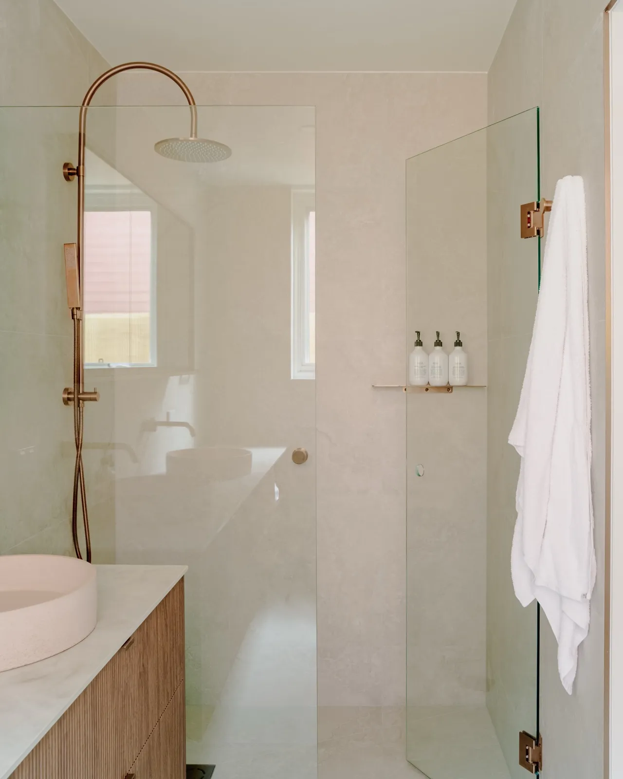 Travertine-look bathroom with brushed copper rainfall shower, glass screen, fluted timber vanity, and round blush vessel basin.