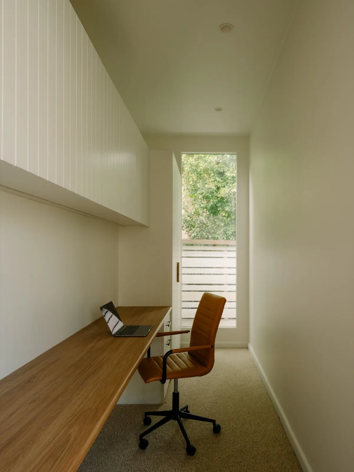 Compact home office with a long timber desk, tan leather chair, and slatted privacy screen at the window.