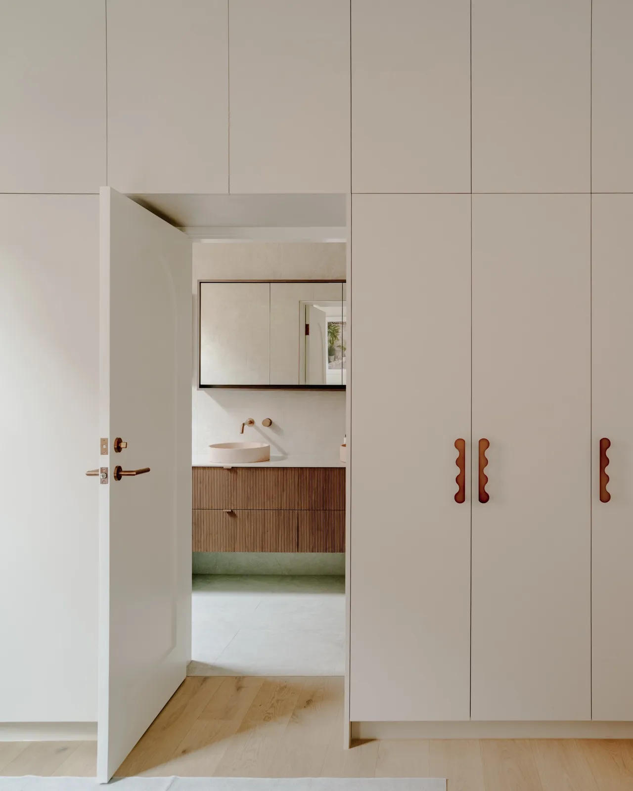 Cream wardrobe wall with wavy timber handles and an open arched door revealing a fluted-timber vanity and round blush basin.