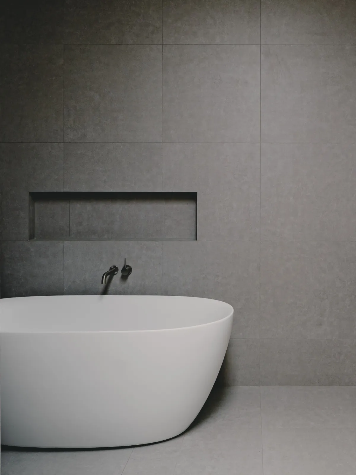 Freestanding tub against a grey large-format tiled wall with a recessed niche and matte-black wall-mounted tap.