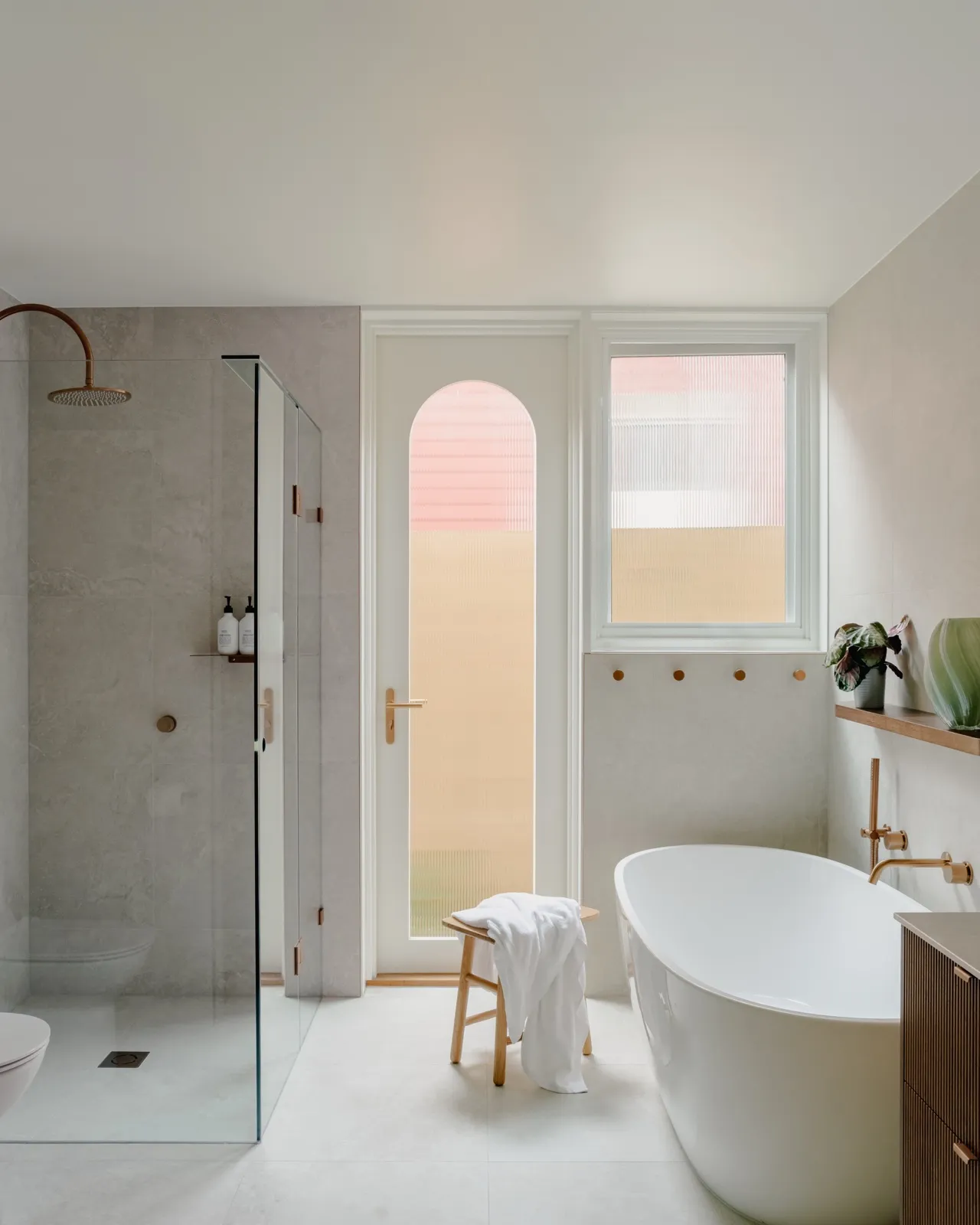 Main bathroom with freestanding tub, walk-in shower, arched glass door, and a window dressed in fluted glass against grey large-format tiles.