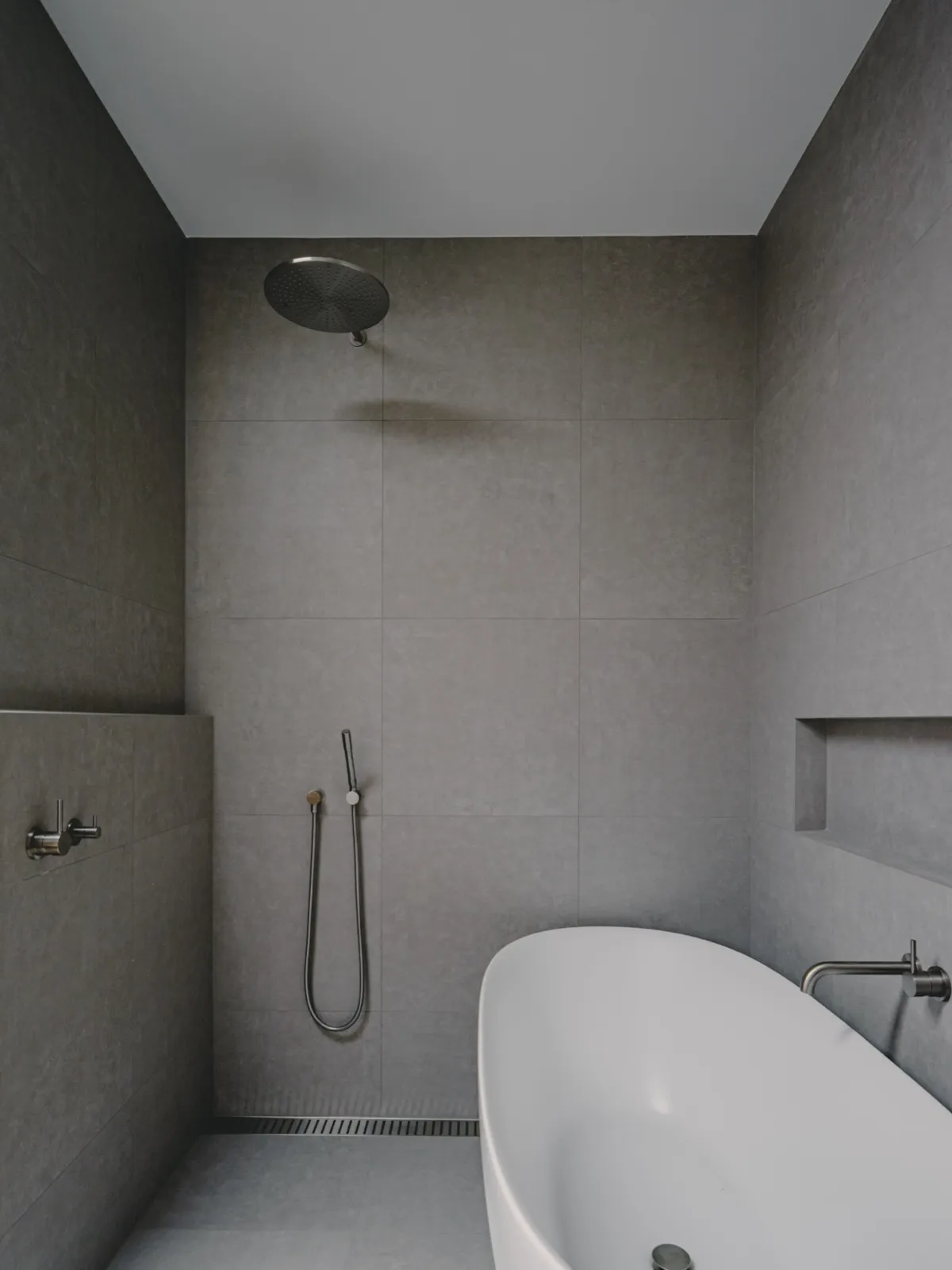 Bathroom with rainfall shower head, hand shower on rail, recessed niche, freestanding tub, and grey large-format tiles.