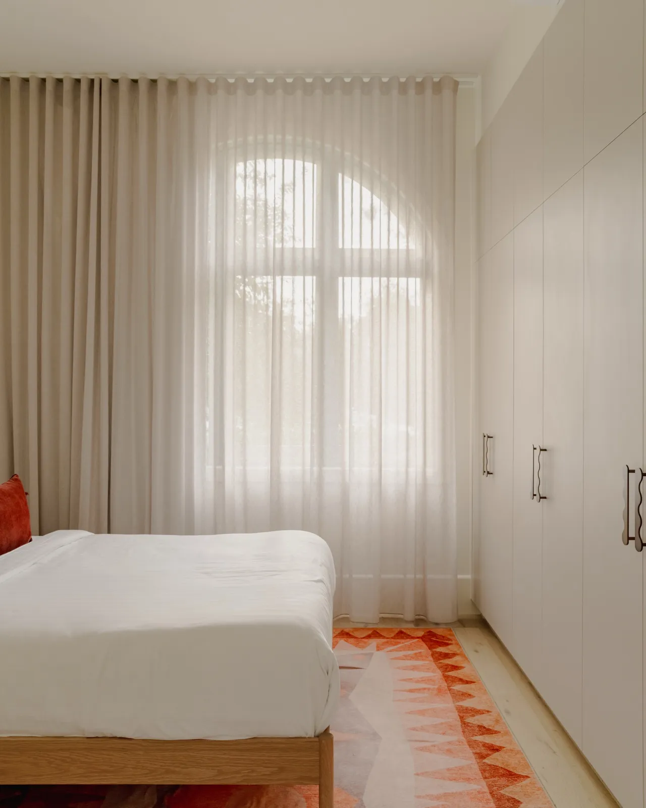 Bedroom with a timber-framed bed, sheer curtains over an arched window, full-height wardrobes, and a patterned orange floor rug.