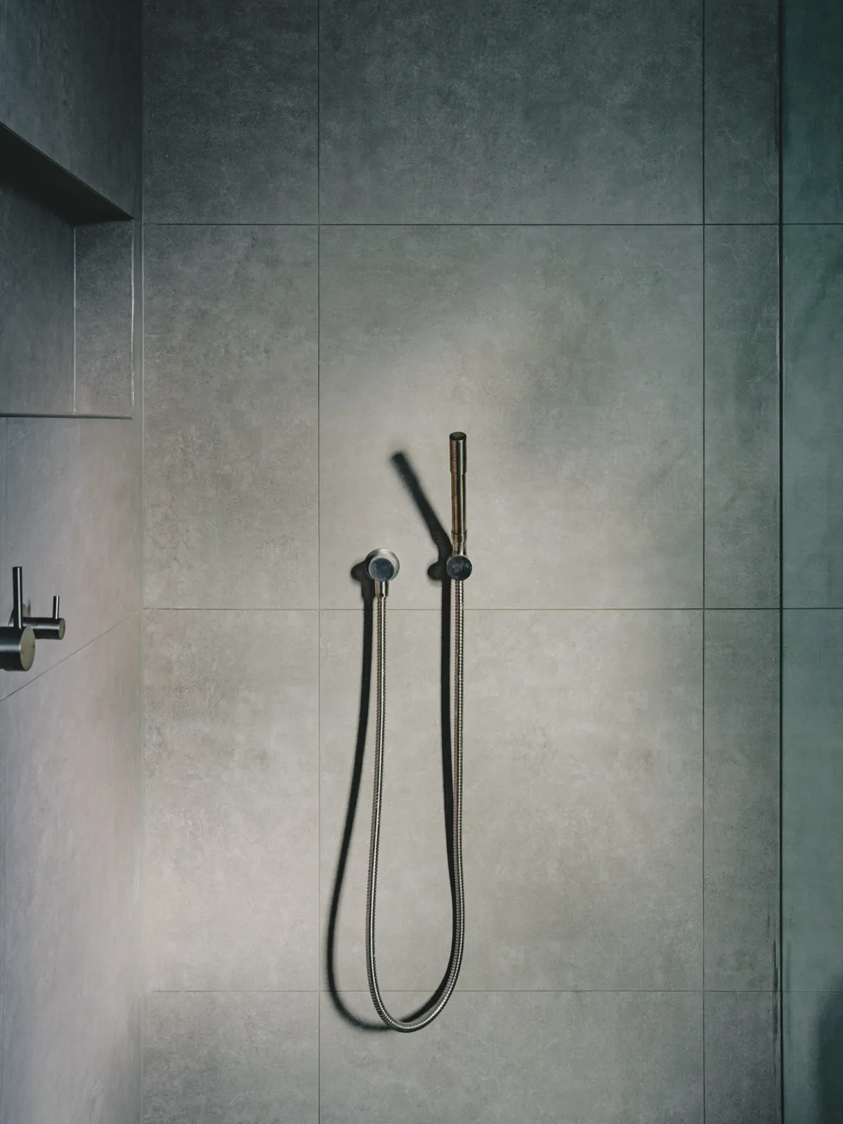 Detail of a brushed gunmetal hand shower on a wall rail against grey large-format tiles.