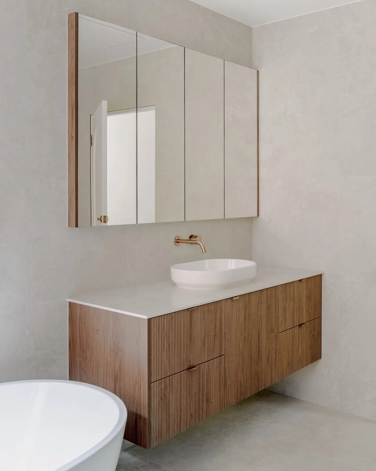Floating fluted timber vanity with a round white vessel basin, brushed copper wall mixer, and a timber-framed mirror cabinet against stone-look tiles.