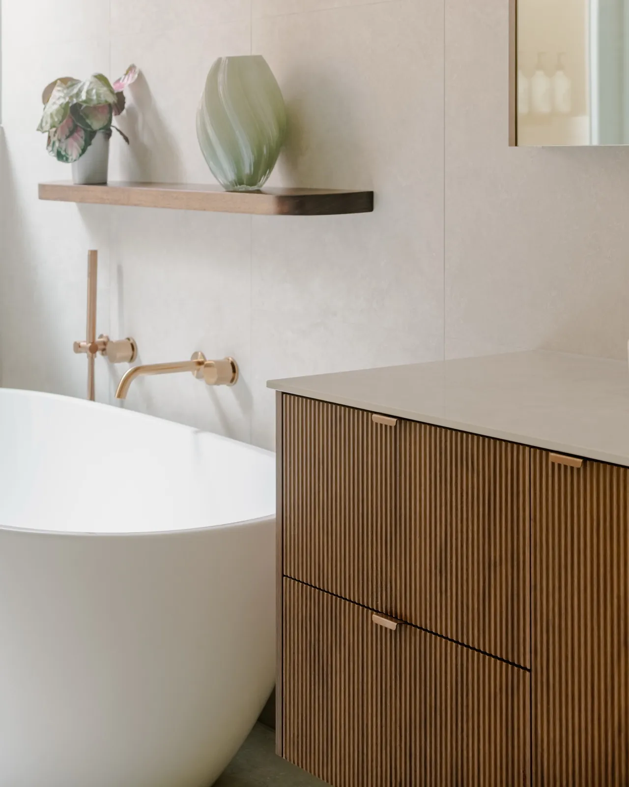 Detail of the freestanding tub with timber floating shelf, sculptural green vase, and a brushed copper wall-mounted bath mixer.