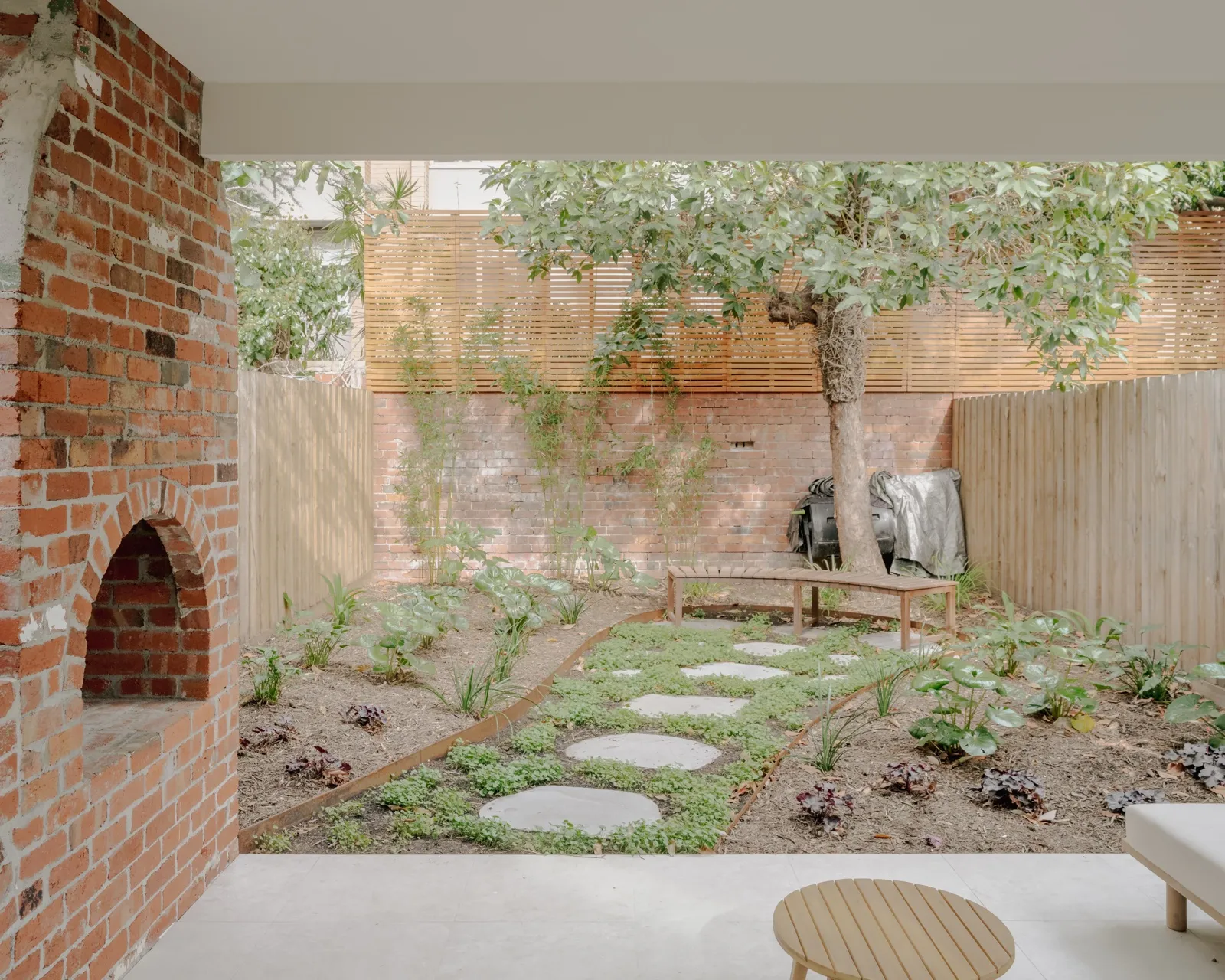 Rear courtyard with retained red-brick chimney, paved stepping-stone path, ground-cover planting, and timber bench seat against the boundary fence.