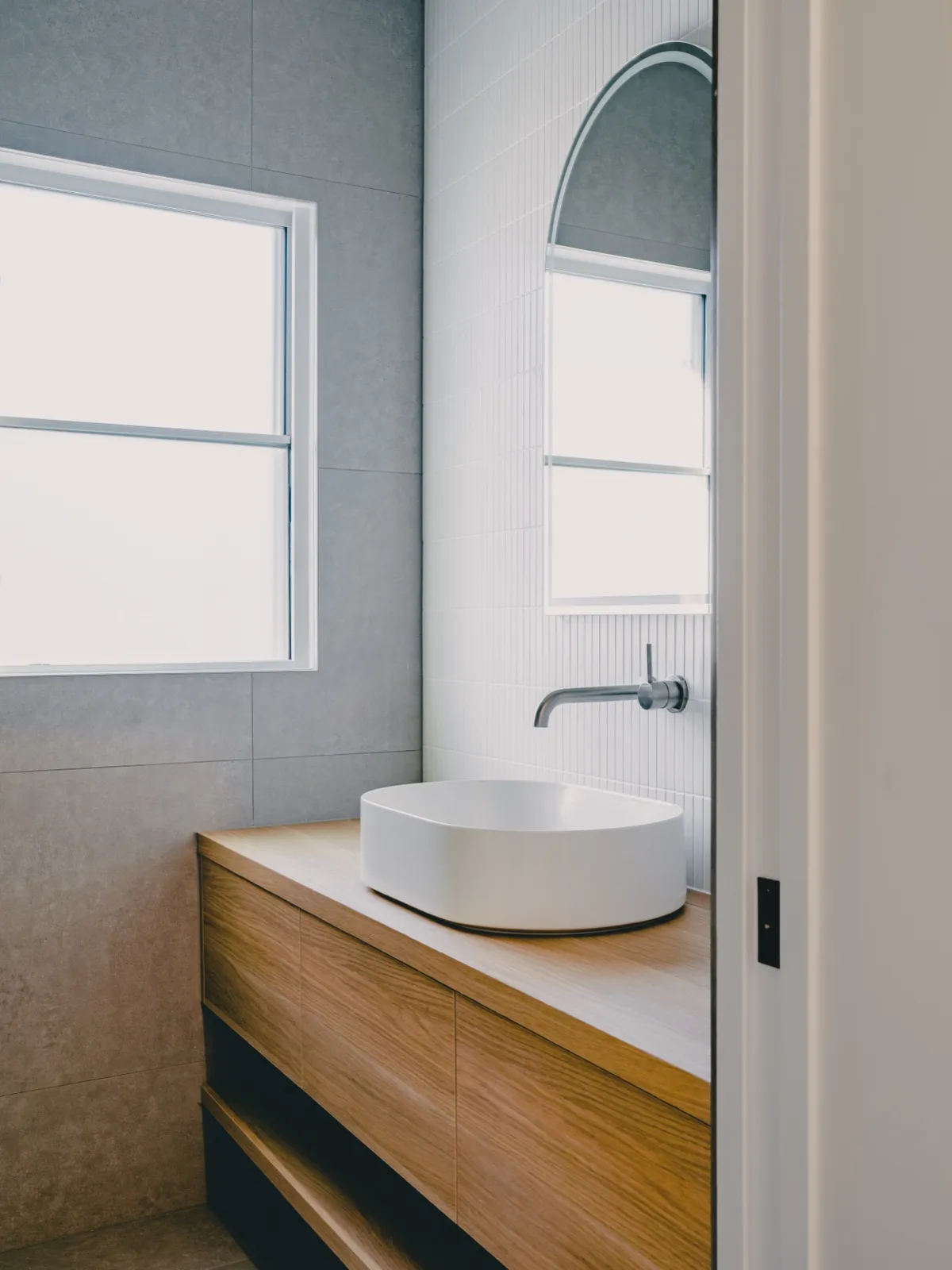 Powder room angled view with grey-tiled walls, arched window, fluted feature wall, and a timber vanity with round basin.