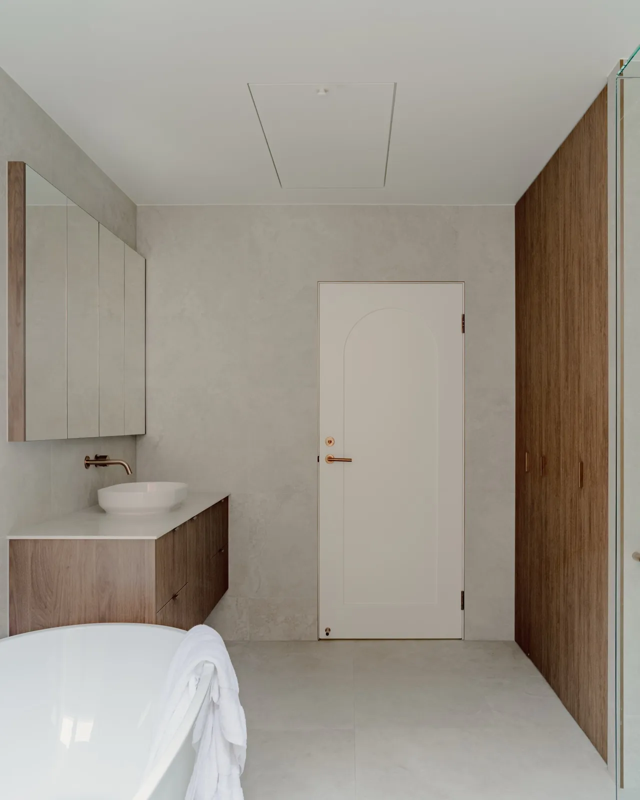 Ensuite with stone-look tiles, freestanding tub, blush wall-hung basin, brushed copper tapware, and an arched solid door.