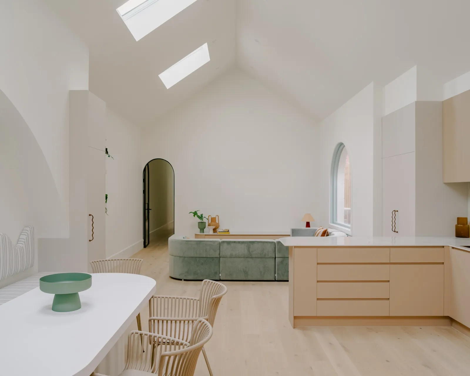 Wide view of the vaulted living room with skylights, arched internal door, blush island, sage sectional, and a white dining table with rattan chairs.