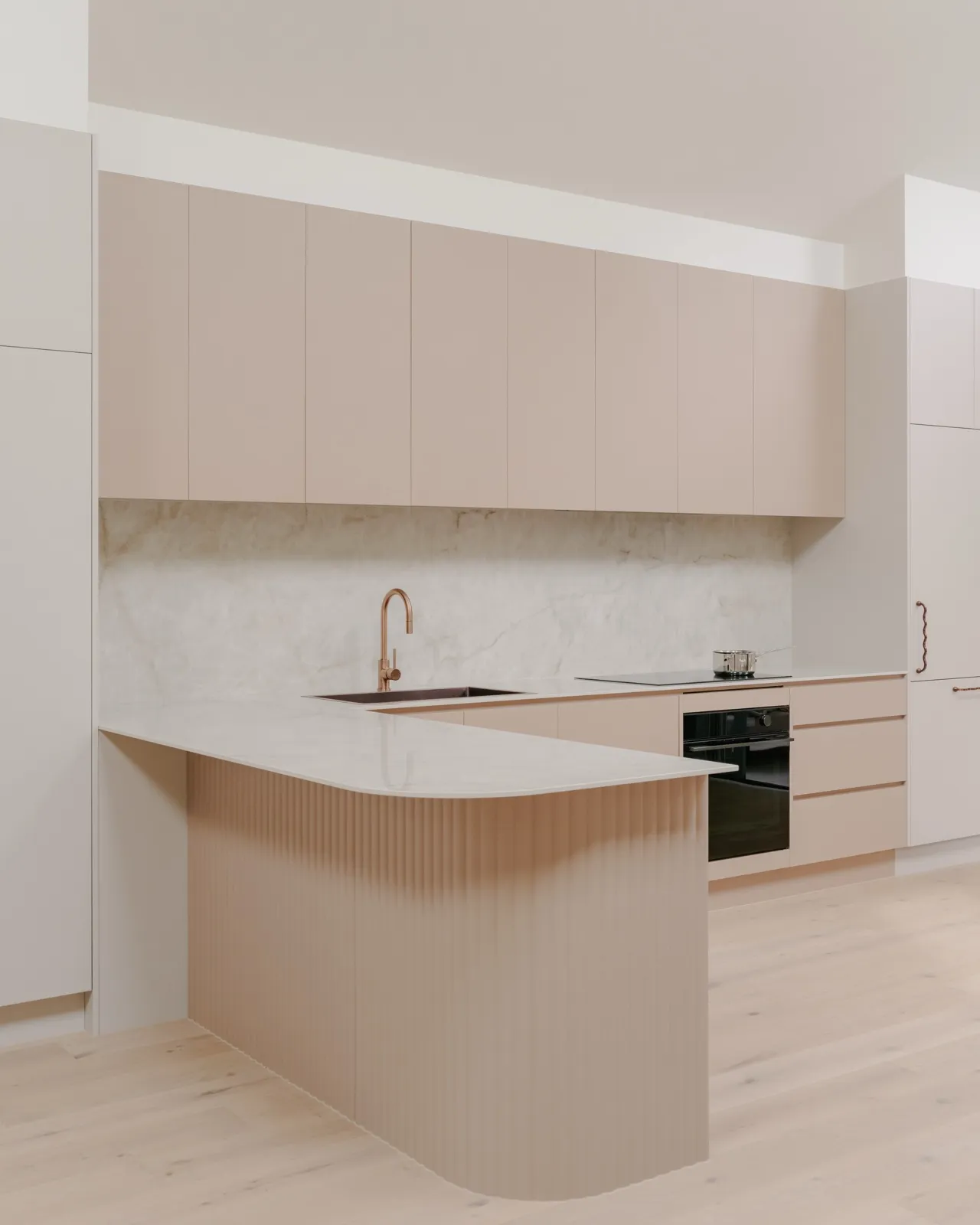 Curved fluted blush island peninsula with white stone benchtop, brushed copper tapware, and a marble splashback behind the kitchen run.