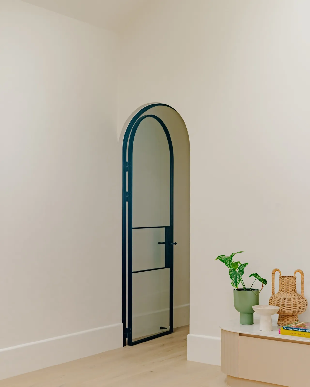 Hallway corner with an arched black-framed internal door and a side console styling green planter, rattan vase, and gardening book.