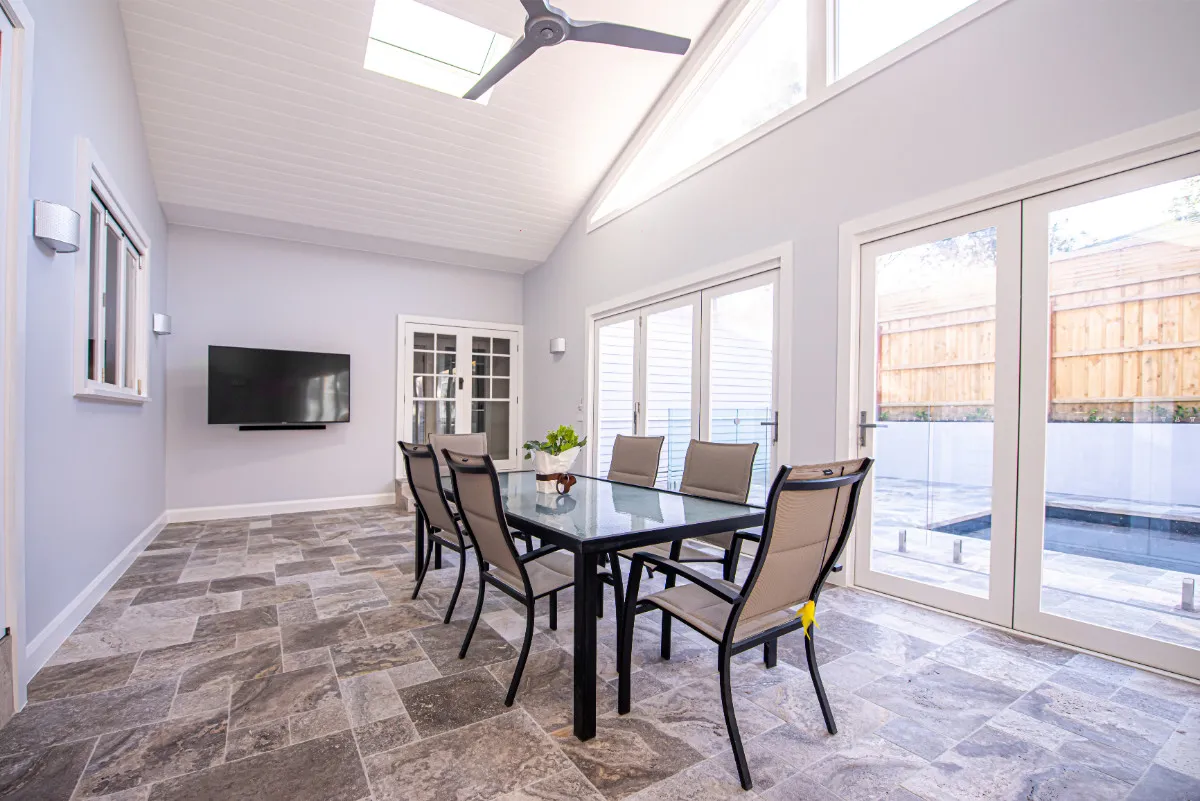 Sunroom alfresco with vaulted ceiling, ceiling fan, glass-top dining setting, and view to swimming pool.