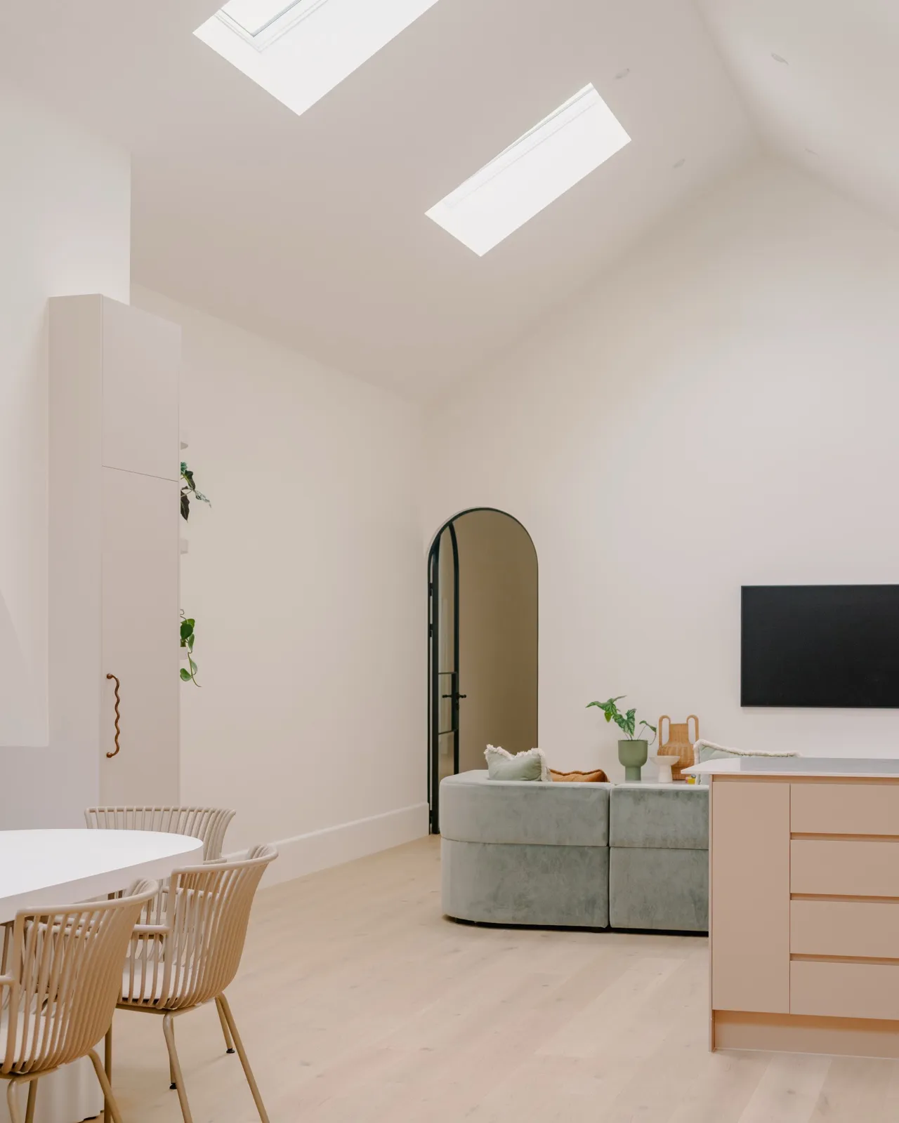 Vaulted living space with arched internal door, sage curved sofa, wall-mounted TV, blush island, and a pale timber floor.