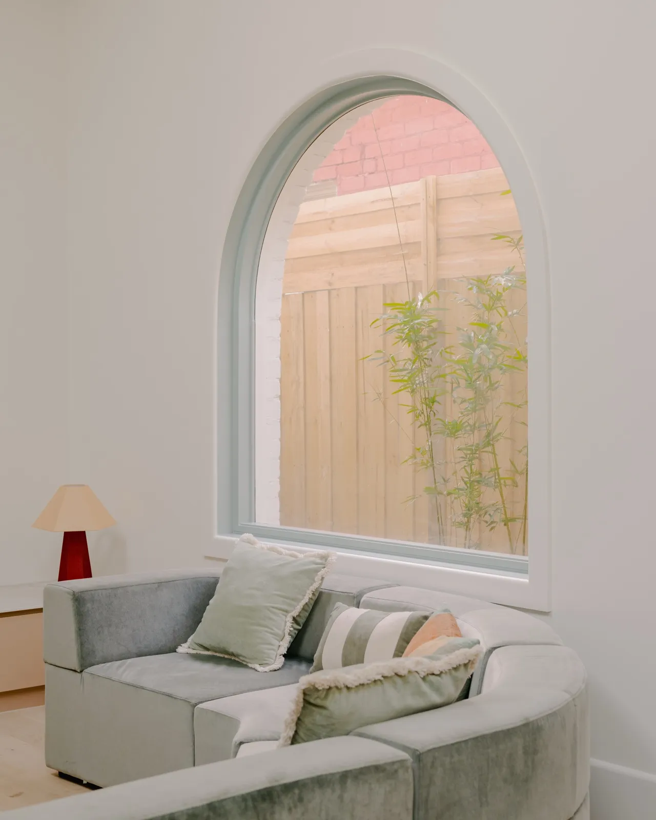 Sitting nook with sage modular sofa, striped and rust cushions, table lamp on cabinet, and an arched window framing a timber boundary fence.