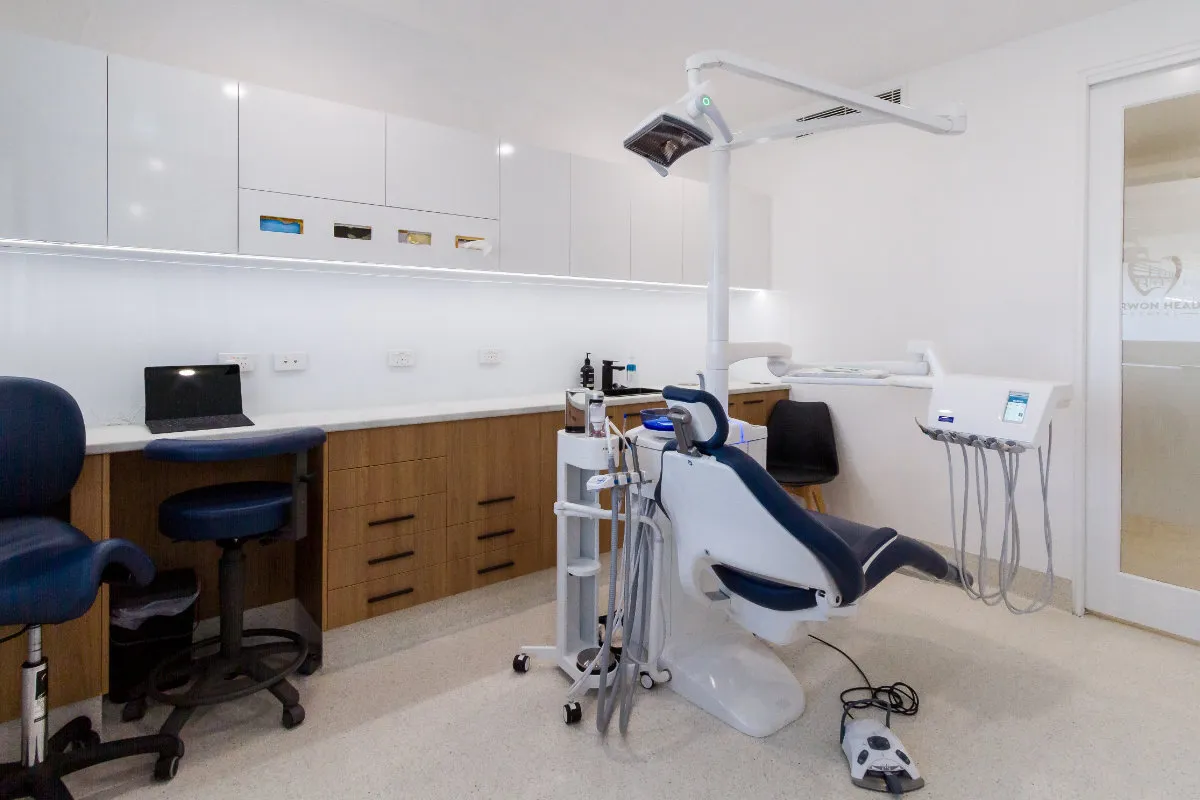 Dental surgery room with chair, overhead light, white joinery, and a coloured-glass feature panel.