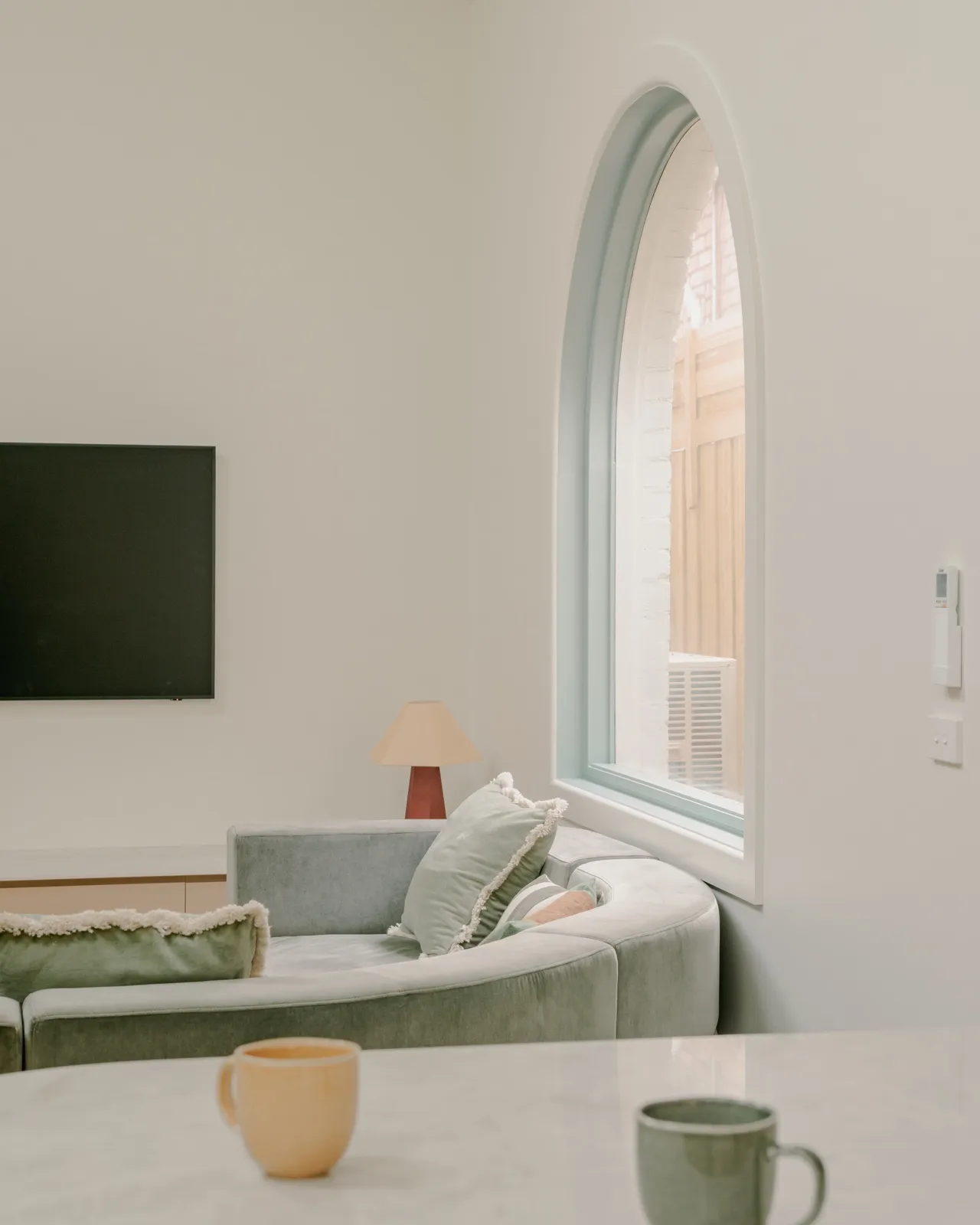 Living corner with curved sage sectional sofa under an arched mint-framed window, table lamp, and wall-mounted TV.