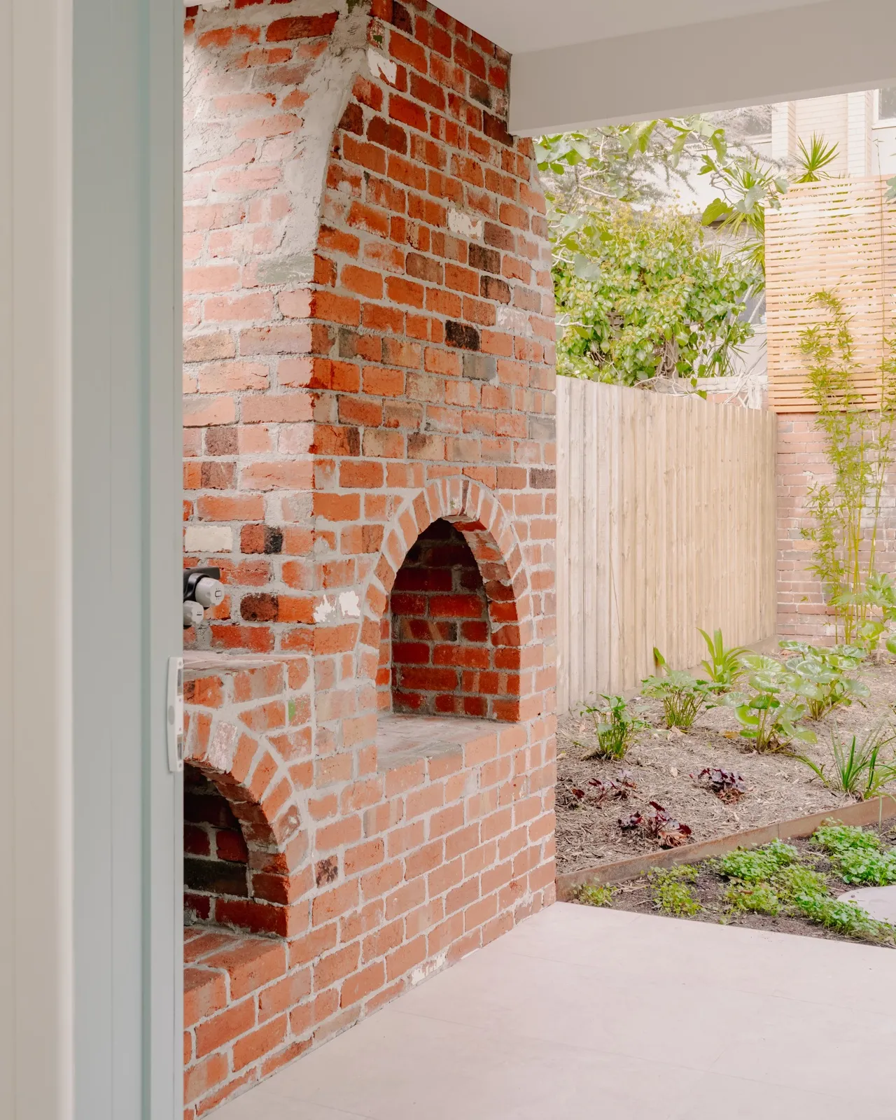 Old red-brick chimney with twin arched openings retained as a courtyard feature beside the rear garden bed and timber fence.