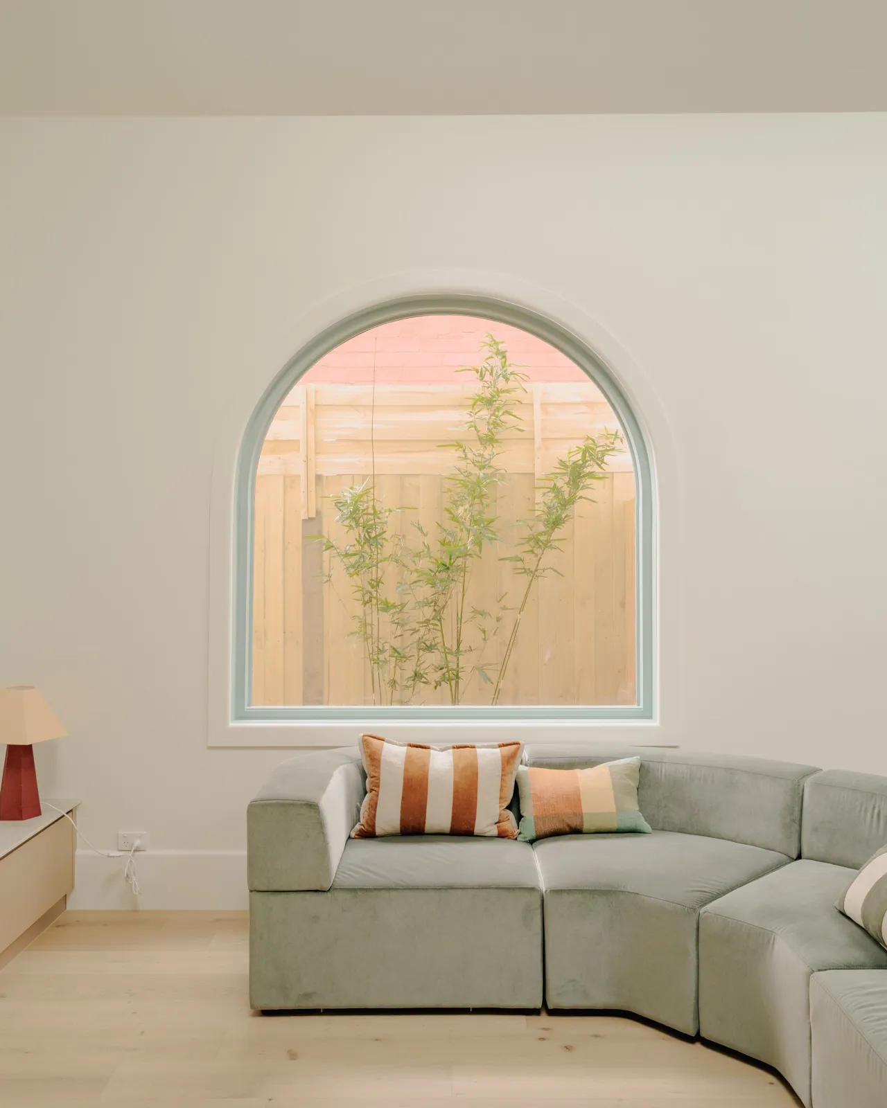Curved sage sectional sofa with rust-striped cushions beneath a large arched mint-framed window over the rear fence.
