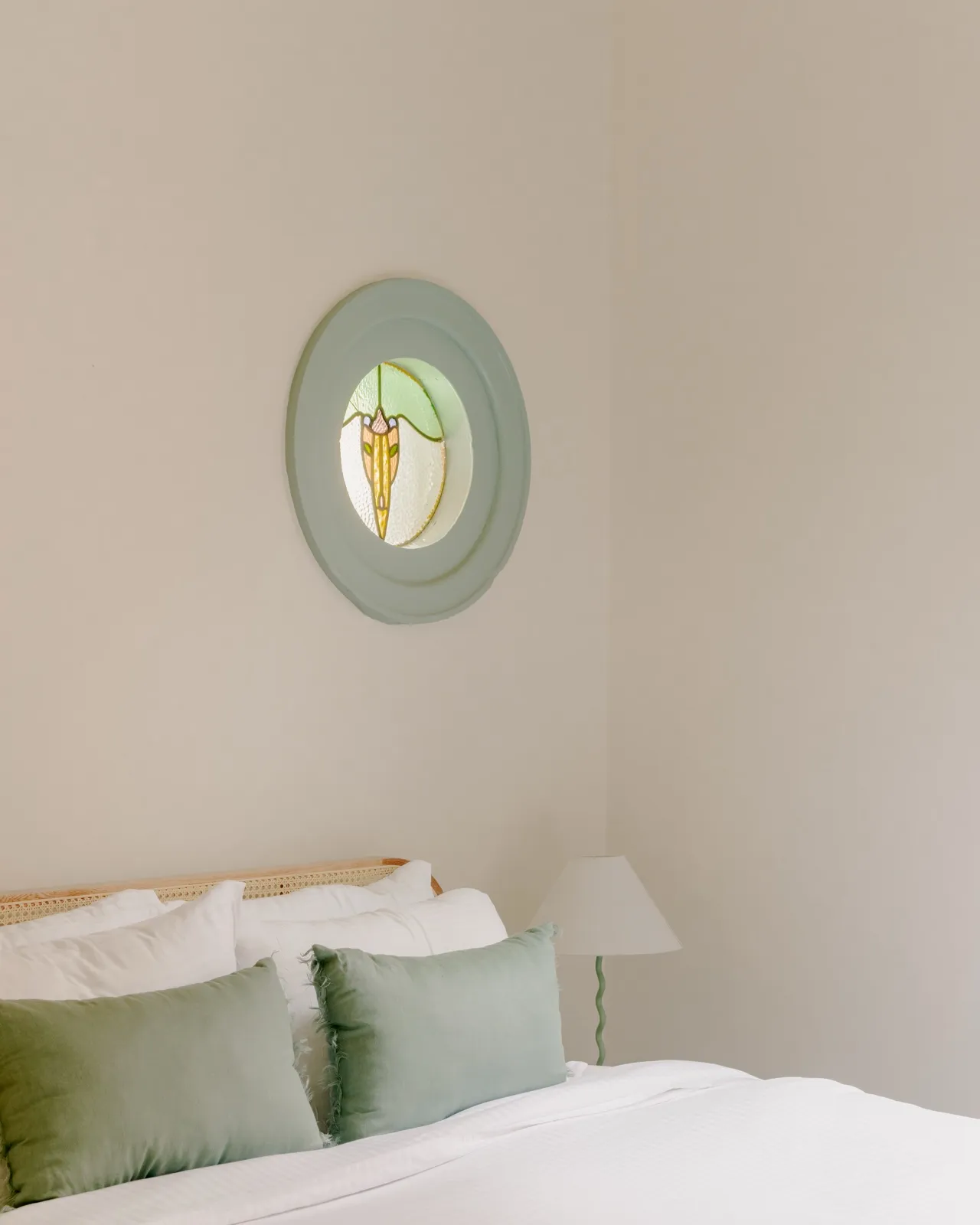 Bedroom detail with a cane bedhead, white linen, sage velvet cushions, and a leadlight stained-glass roundel set into the wall.