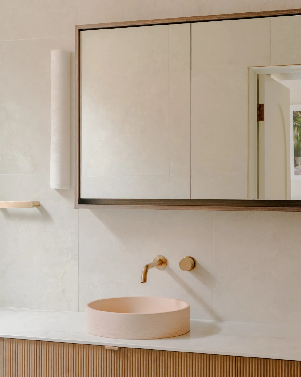 Bathroom detail with blush round vessel basin, brushed copper wall mixer, and a timber-framed mirrored cabinet against stone-look tiles.