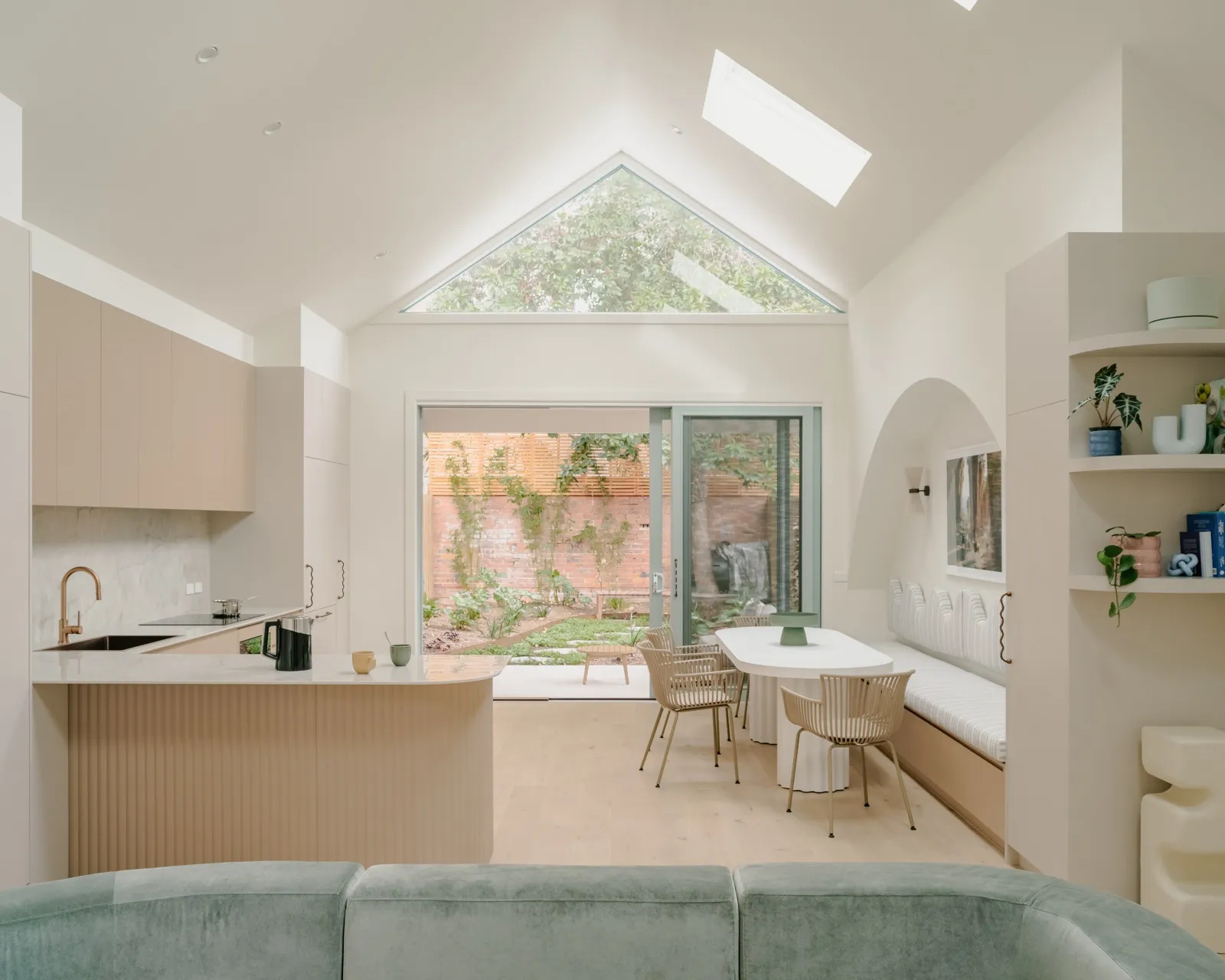 Wide rear living view with pitched ceiling, fluted blush island, sage sofa, banquette dining, and sliding doors to the courtyard garden.