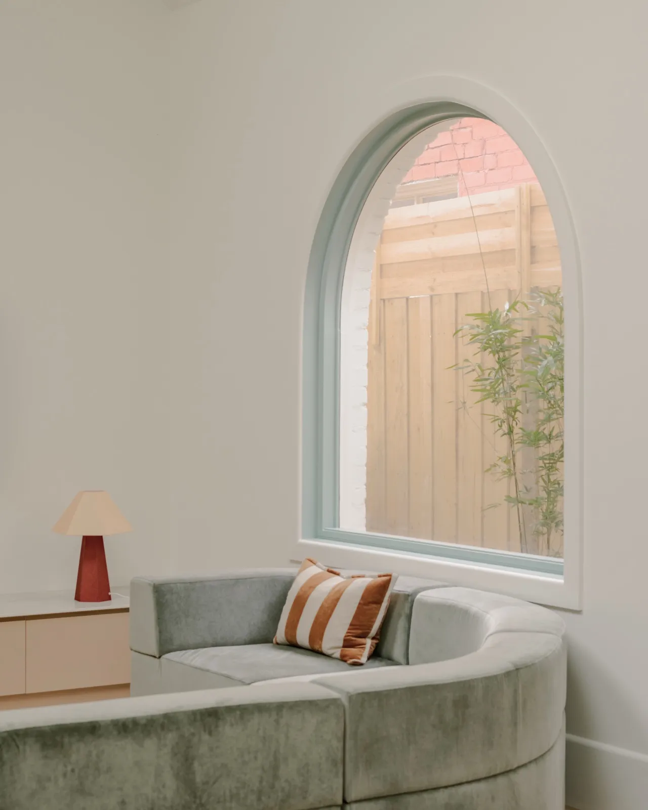 Sage sectional sofa beside a console with a red and tan table lamp under an arched mint-framed window onto the timber fence.