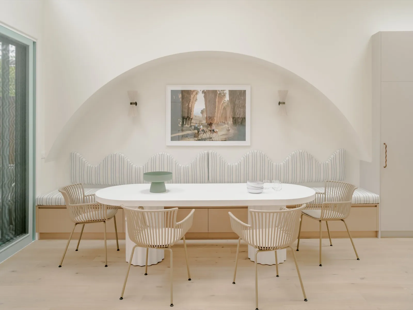 Dining alcove with arched plaster recess, framed Italian-cypress artwork, twin pleated sconces, and a white pedestal table with rattan armchairs.