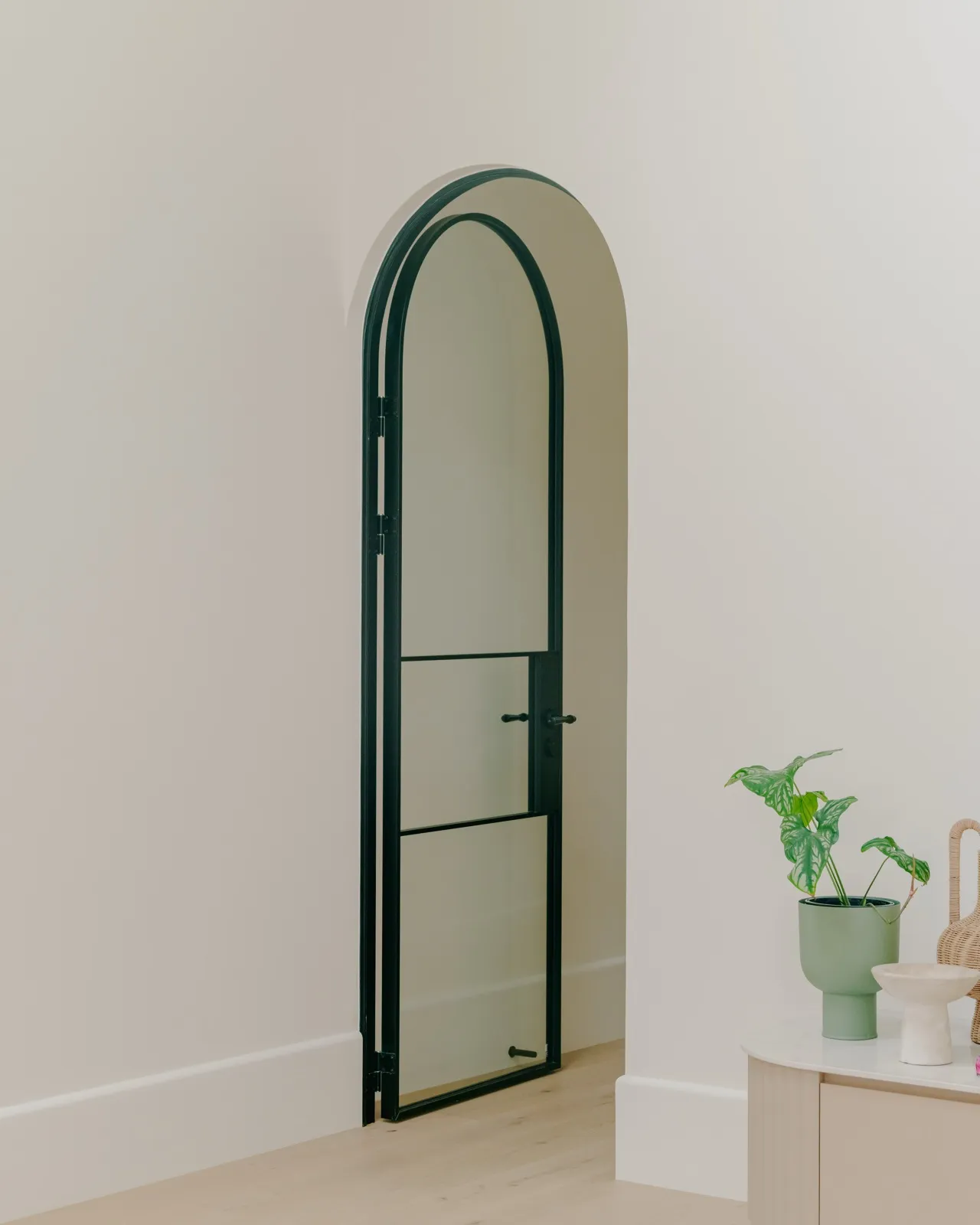 Black-framed arched internal door closed against a white wall, with a styled blush console and green planter to the side.