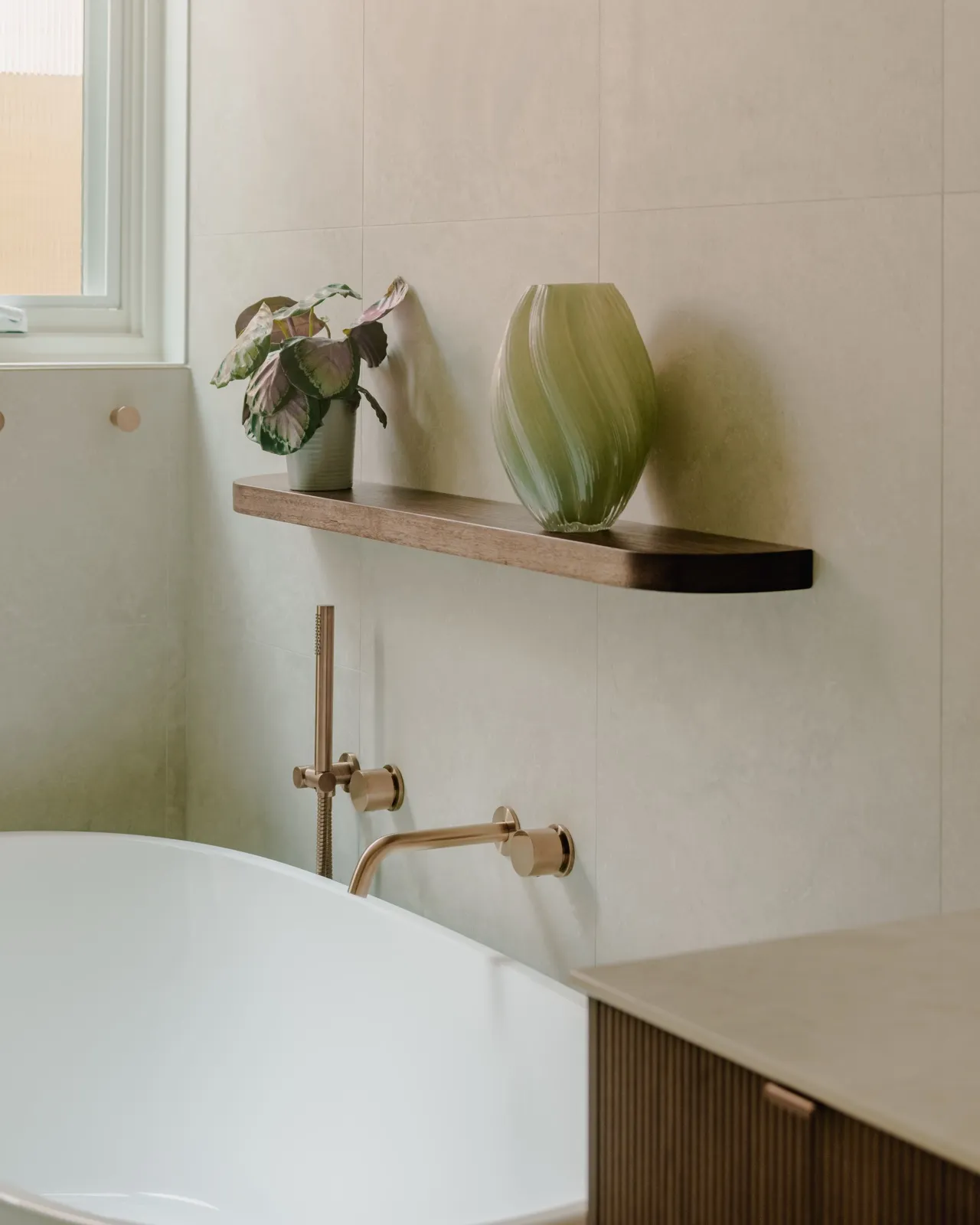 Bath detail with brushed copper hand shower and wall mixer, timber floating shelf, leafy plant, and a green art-glass vase.