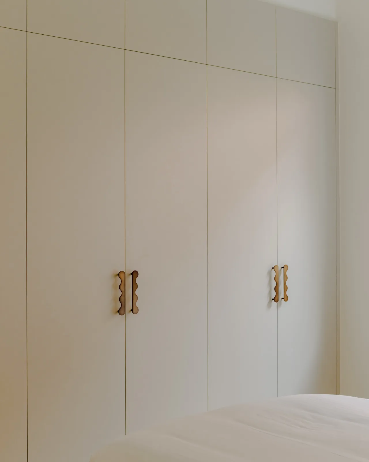 Built-in cream wardrobe doors with sculptural wavy timber pull handles beside a made bed with white linen.